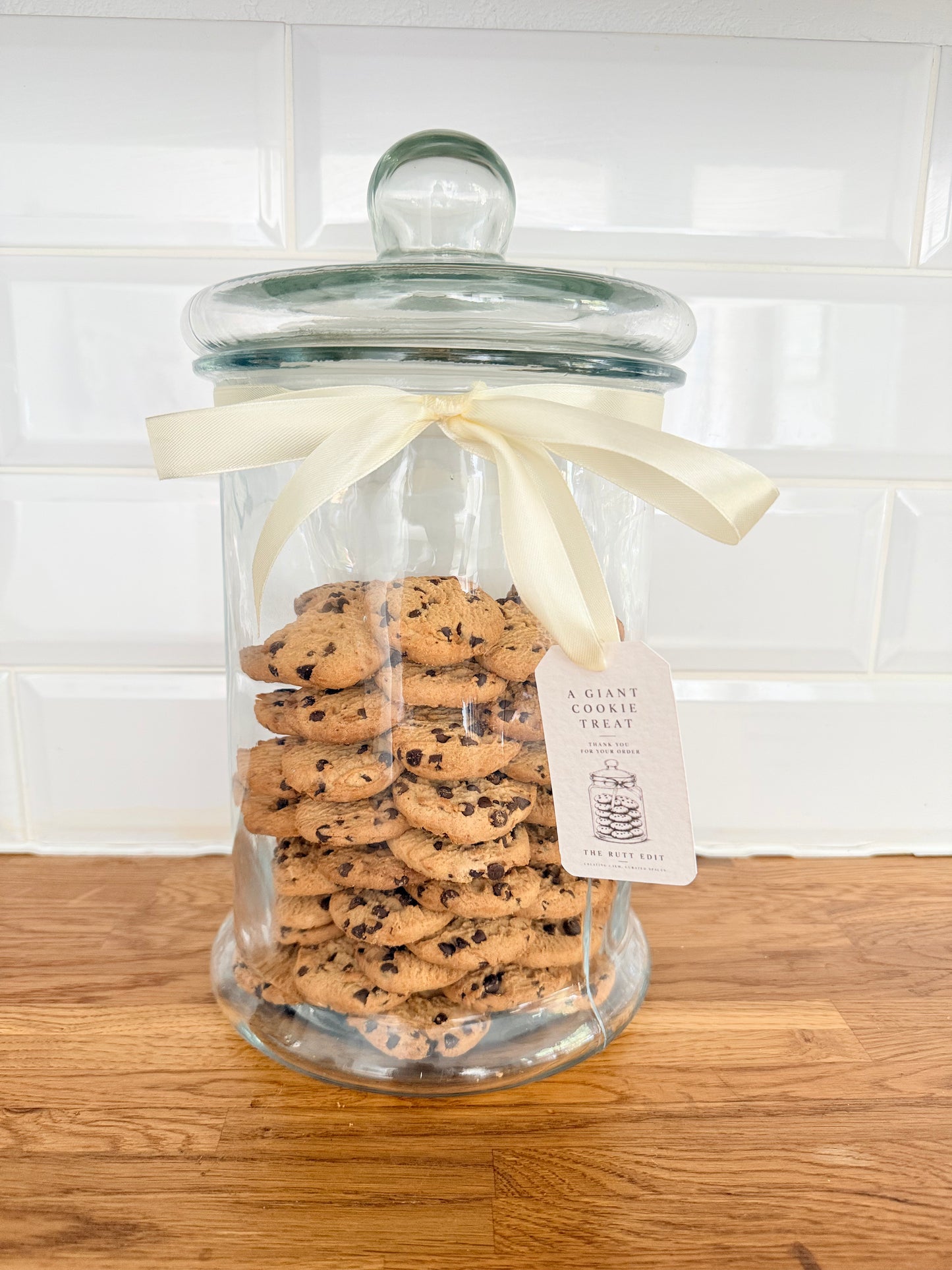 A large clear glass jar filled with assorted chocolate chip cookies, topped with a cream ribbon bow and a hanging paper tag, set on a wooden surface against a white tiled wall.
