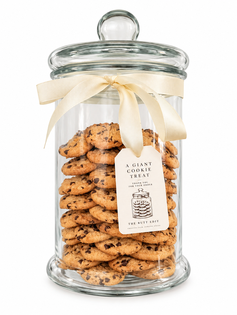 A clear glass jar filled with chocolate chip cookies, tied with a cream ribbon and featuring a paper tag that reads 'A GIANT COOKIE TREAT'