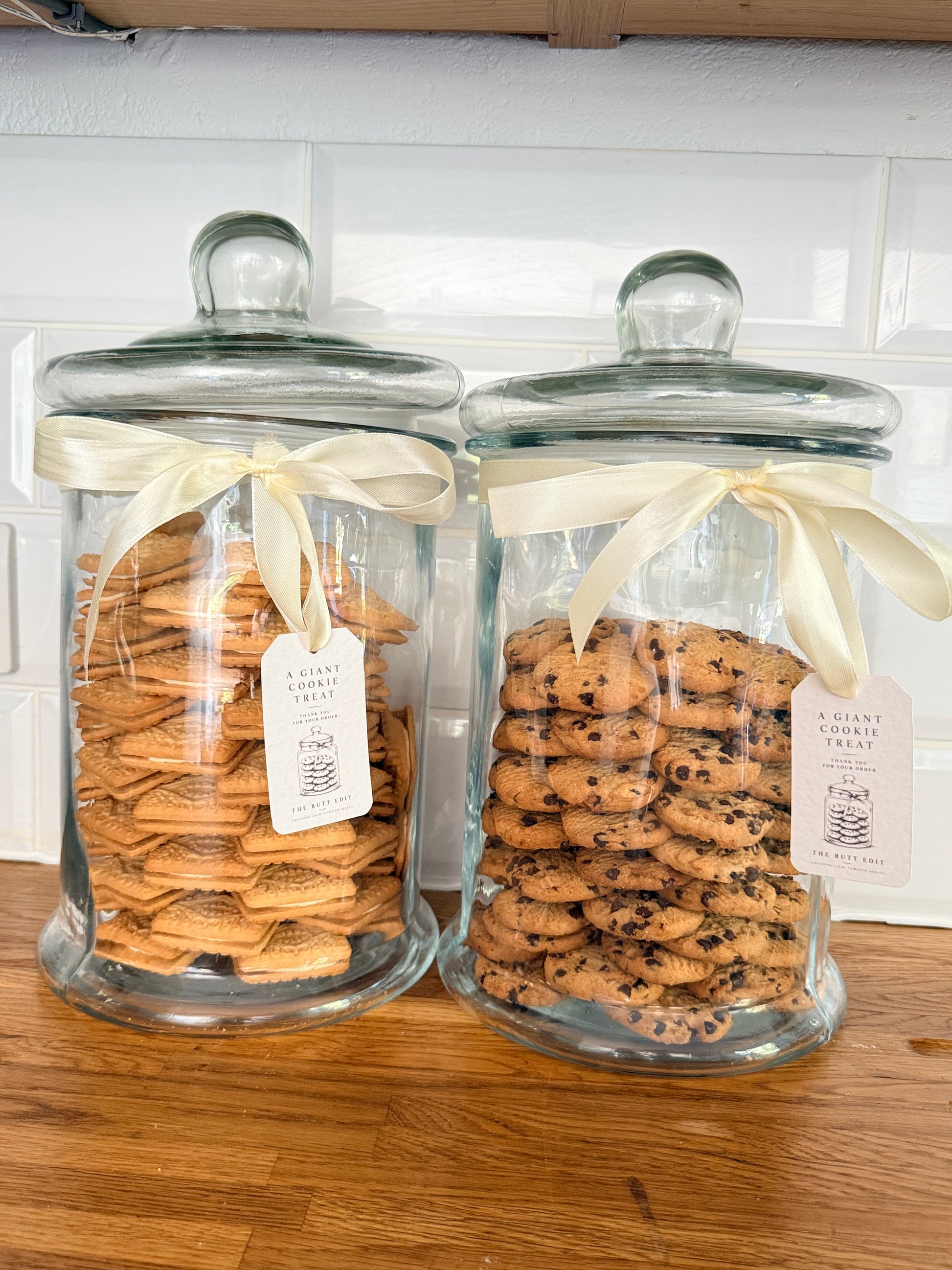 Two Giant Cookie glass Jars with lids, tied with cream ribbons, filled with assorted cookies on a wooden countertop against a white tiled wall. One canister contains golden-brown biscuits and the other chocolate chip cookies.